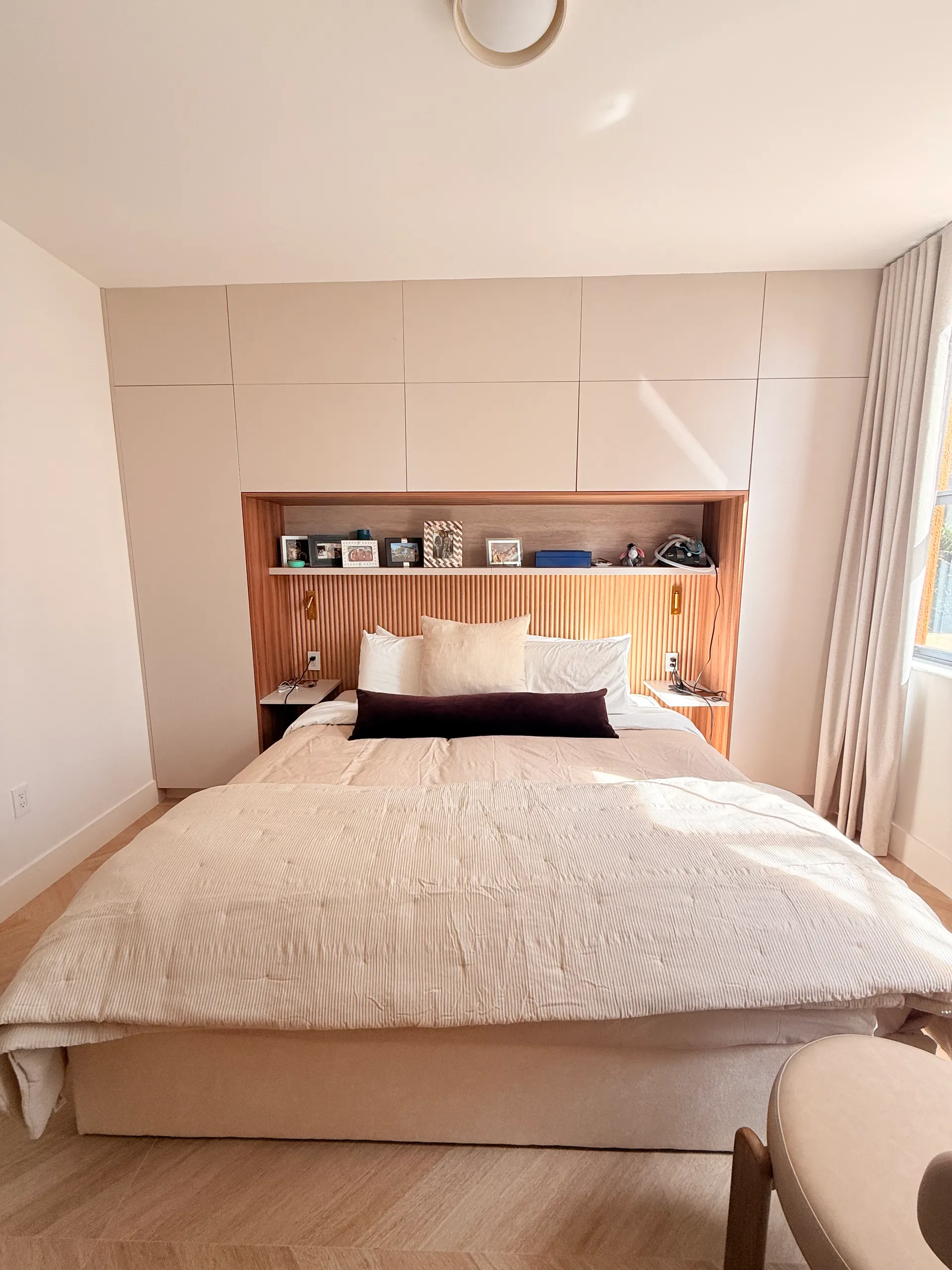 El dormitorio terminado: la cama contra el cabecero acanalado de roble, un estante iluminado con fotos encima y mesas de noche flotantes a ambos lados