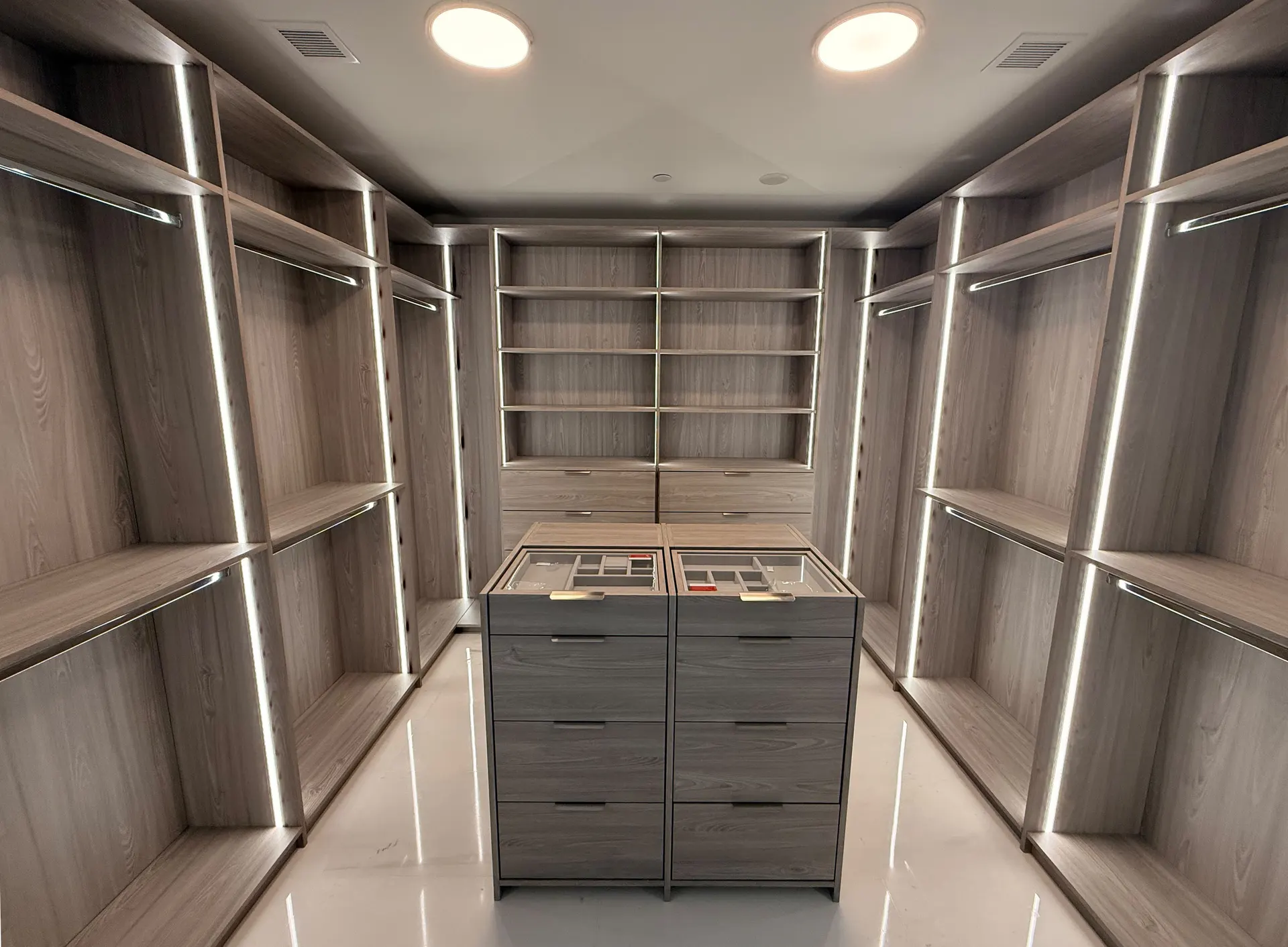 Walk-in closet en roble gris con tubos iluminados a ambos lados y una isla central de gavetas con bandejas de joyería con tapa de vidrio