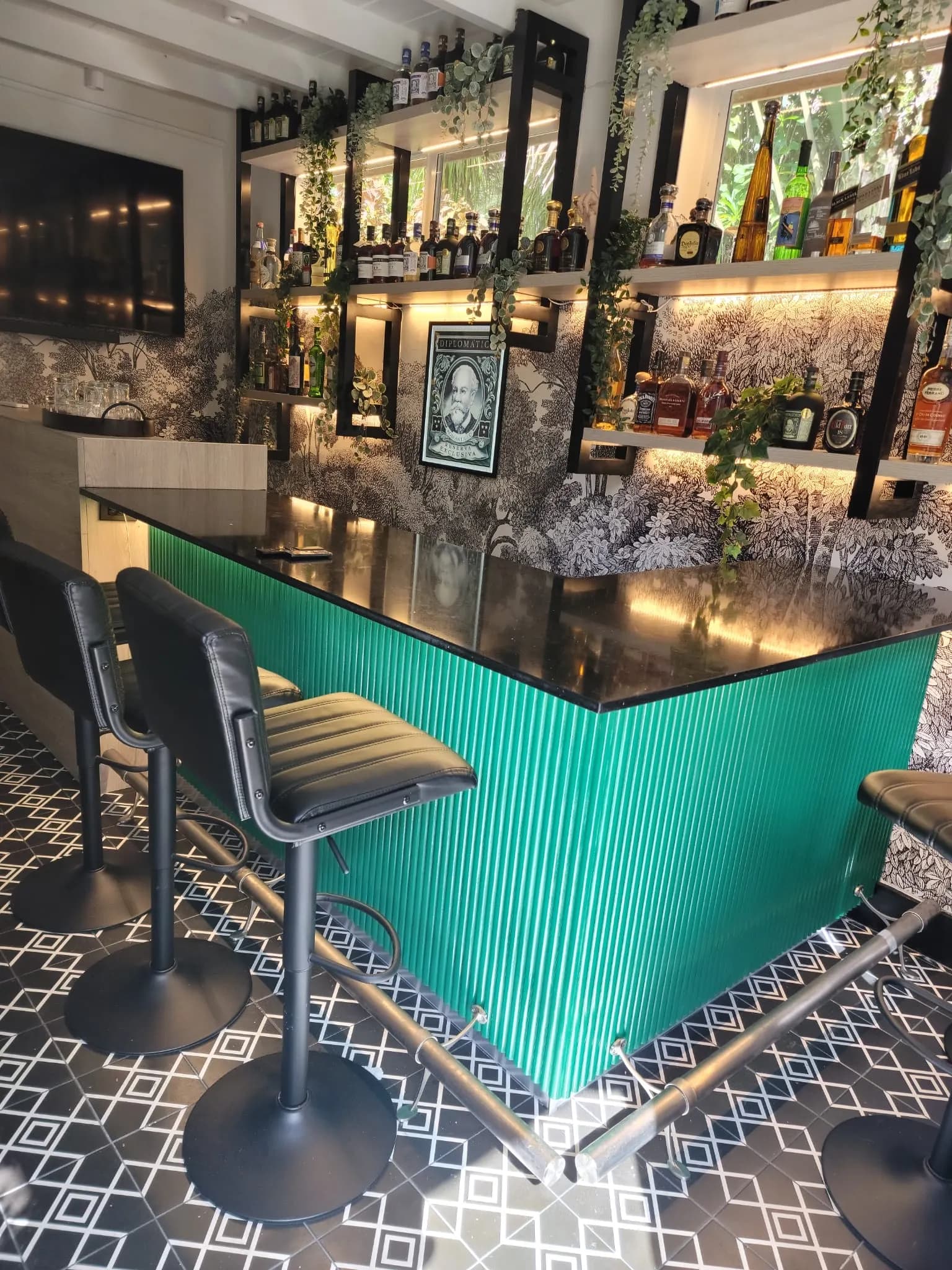Barra a medida con frente acanalado verde, tope de piedra negra y estantería retroiluminada con botellas y plantas colgantes
