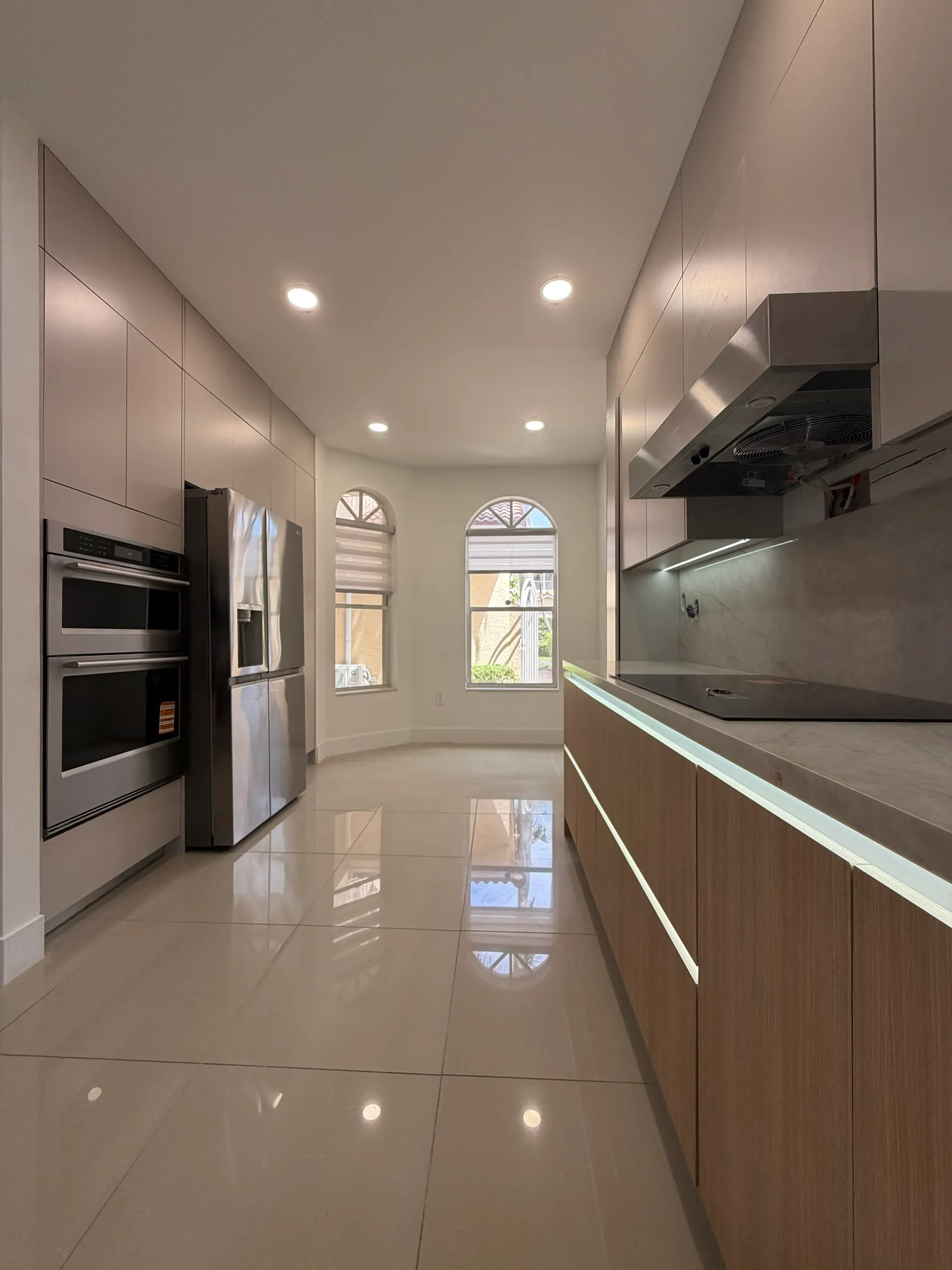 Cocina tipo pasillo con una línea de gabinetes bajos de nogal, tope de mármol con luz bajo el borde, campana de acero sobre la hornilla y una pared de gabinetes sin tiradores alrededor del horno doble y la nevera empotrados