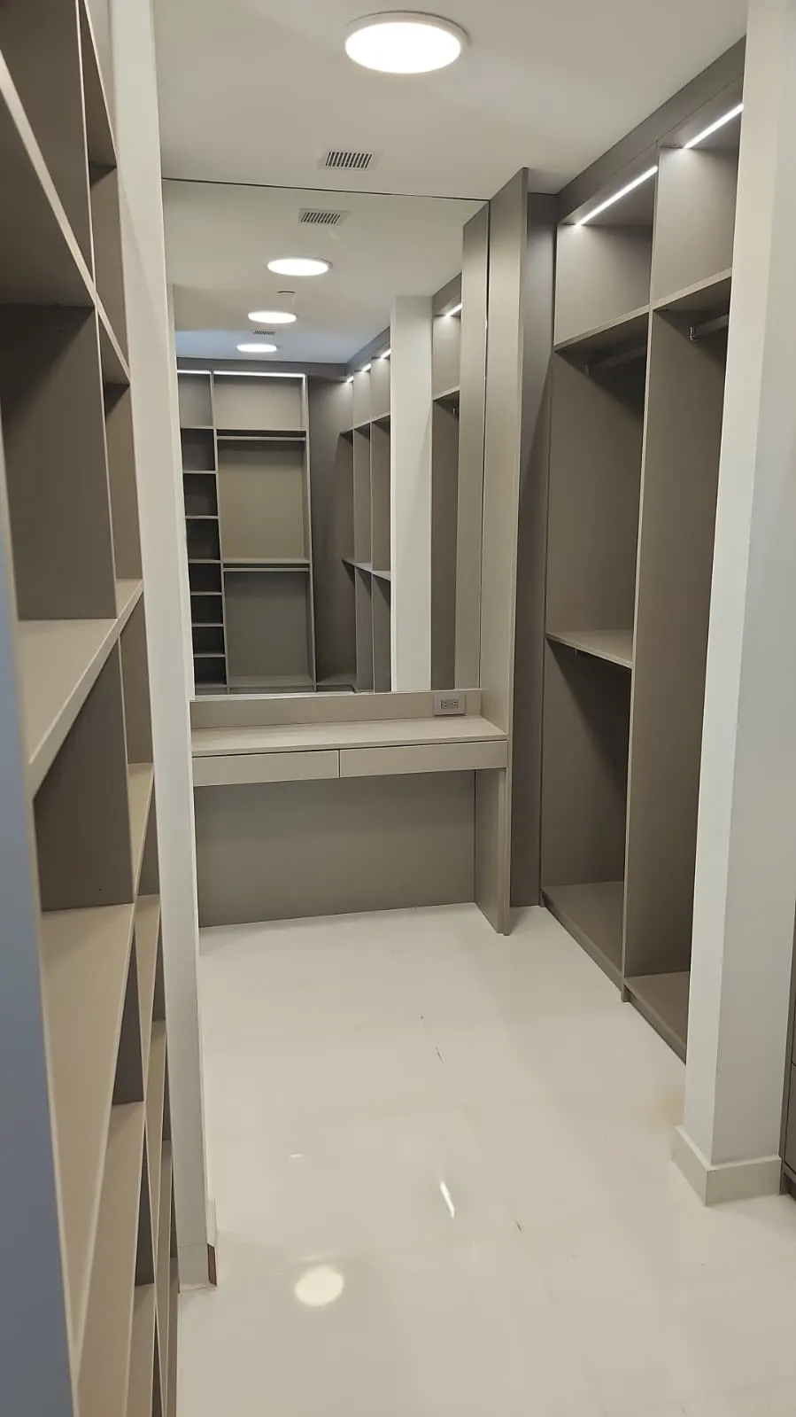 Walk-in closet en gris arena con estantes y tubos a ambos lados, tiras LED en lo alto y un tocador con espejo amplio al fondo